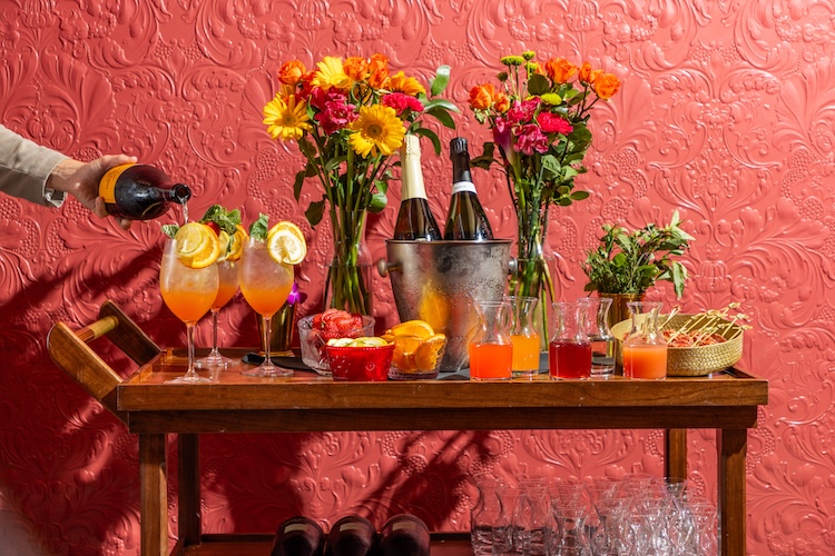 A spritz cart full of drink ingredients and vases full of flowers against a salmon-colored wall.