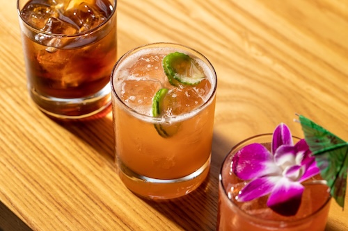 A lineup of cocktails on a wooden surface, one dark amber and one orange-hued with lime and purple orchid garnish